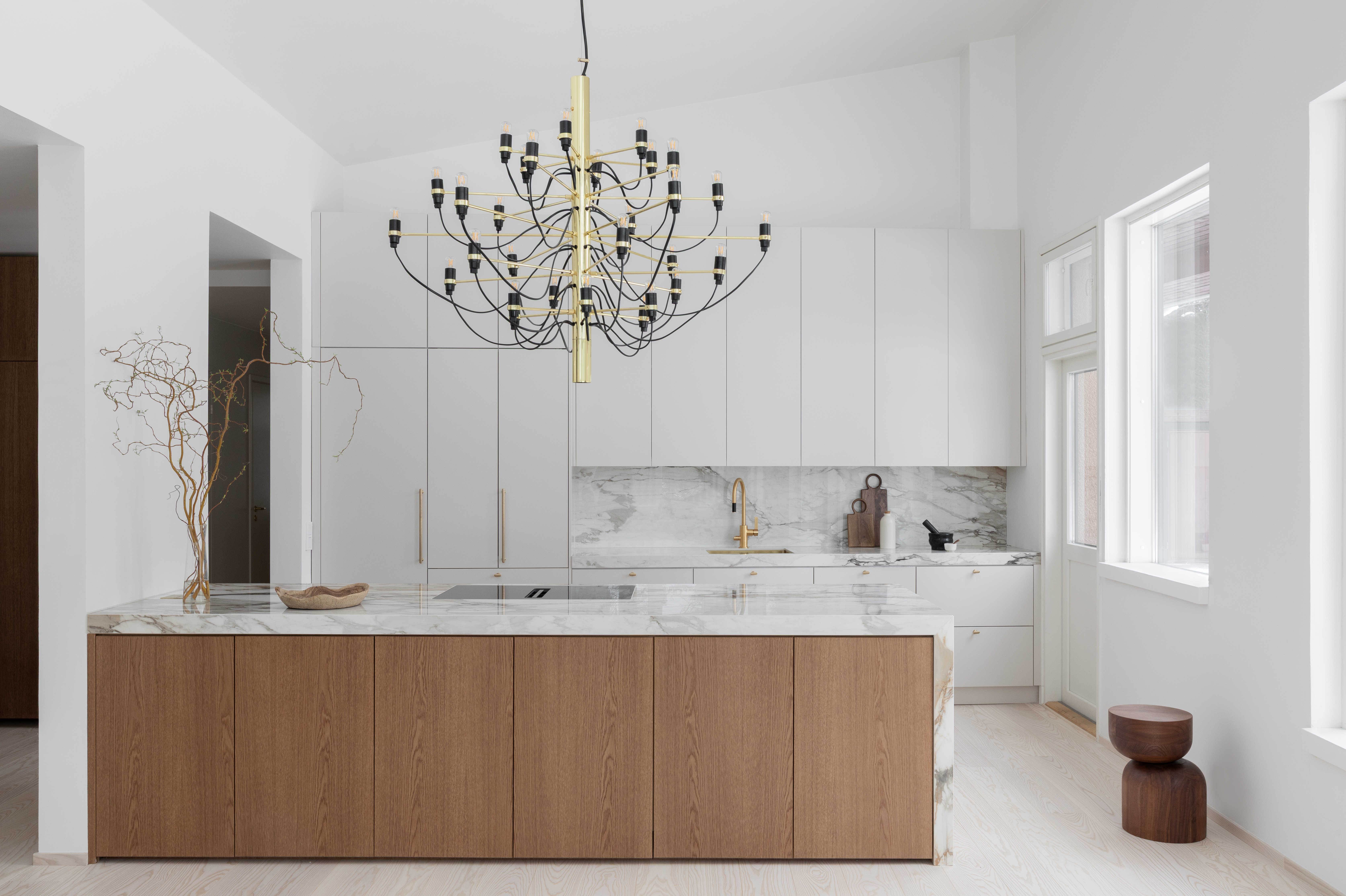 Moderni designkeittiö, jossa hiekan sävyinen mulperi-maalattu kaapisto, marmorikuvioinen keraaminen työtaso ja välitila sekä tummasta tammiviilusta tehty keittiösaareke.